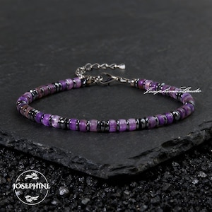 Puede incluir: Una pulsera de cuentas moradas y negras con detalles metálicos plateados. La pulsera se muestra sobre una superficie oscura. El texto "Josephine Studio" es visible en la pulsera.