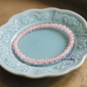May include: A delicate pink beaded bracelet rests on a light blue, ornate plate. The bracelet is composed of small, round, rose quartz beads. The plate has a floral design around the rim. The image has a soft, natural lighting.