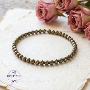 May include: A beaded bracelet made of small, round, golden-colored beads. The bracelet is displayed on a textured, off-white surface, with dried pink roses in the background. The bracelet is a jewelry item.