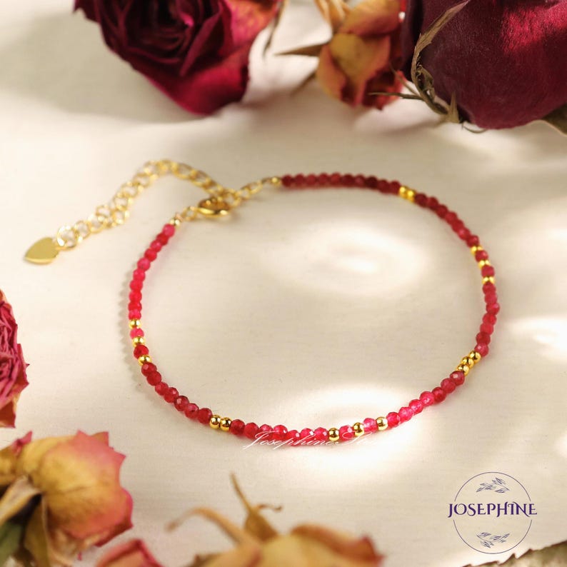 Pu&ograve; includere: Un delicato braccialetto a catena dorata con un ciondolo a forma di cuore e perline di pietre preziose rosse.