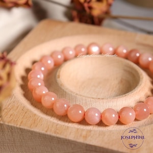May include: A peach-colored sunstone bracelet with a red string closure. The bracelet is on a wooden surface.