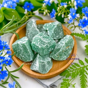 May include: A wooden bowl filled with green aventurine tumbled stones. The stones are smooth and polished, with a light green color and white flecks. The bowl is surrounded by green leaves and blue flowers.