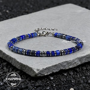 Puede incluir: Una pulsera de cuentas azules oscuras y plateadas. La pulsera está hecha de pequeñas cuentas azules cuadradas y cuentas plateadas metálicas. La pulsera se muestra sobre una superficie de piedra gris. La pulsera tiene un cierre plateado. La marca "Josephine Studio" es visible.