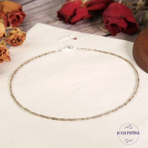 May include: A delicate silver chain necklace with small, light brown beads. The necklace is on a white surface with dried roses in the background.