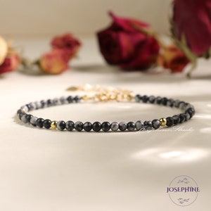 May include: A delicate bracelet made with small black and white marbled beads and gold accents. The bracelet is on a white surface.