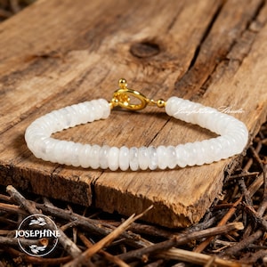 Könnte beinhalten: Ein weißes Perlenarmband mit einem goldenen Verschluss, das auf einer Holzoberfläche liegt. Das Armband besteht aus kleinen, runden, weißen Perlen. Der Verschluss ist ein goldfarbener Knebelverschluss. Das Armband ist mit "Josephine Studio" beschriftet.