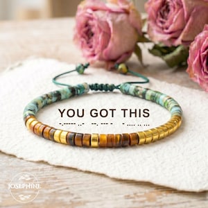 May include: A beaded bracelet with turquoise, brown, and gold-colored beads. The bracelet has the text "YOU GOT THIS" and a Morse code message. The bracelet is on a white surface with pink roses in the background.