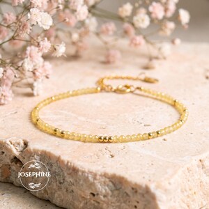 May include: A delicate bracelet featuring small, faceted yellow beads interspersed with tiny, polished gold-tone metal spheres. The piece includes a gold-tone chain extension and a clasp closure. A circular logo with the text JOSEPHINE is visible.