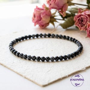 May include: A black snowflake obsidian beaded bracelet. The bracelet is made of small, round, black beads with white snowflake-like patterns. The bracelet is displayed on a textured, off-white surface, with dried pink roses in the background.