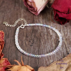 May include: A delicate bracelet featuring iridescent, clear beads strung on a silver chain. The bracelet is displayed on a wooden surface with dried leaves and roses in the background. The bracelet has a small heart-shaped charm and a clasp. The word "JOSEPHINE" is visible.
