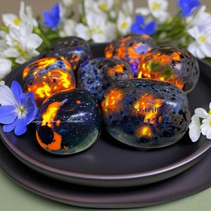 May include: A collection of seven tumbled stones with a black, blue, and orange marbled pattern. The stones are arranged on a black plate. The text "JOSEPHINE" is visible on one of the stones.