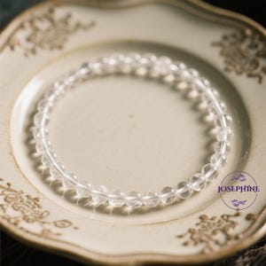 May include: A clear quartz crystal bracelet with round beads on a white plate with a brown floral design. The bracelet is made of natural stones and is a popular choice for jewelry.
