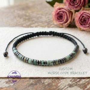 May include: A custom Morse code bracelet with a black braided cord and adjustable closure. The bracelet features a row of small, light green and gray beads, interspersed with dark gray metal beads. The word "JOSEPHINE" is printed on the lower left.