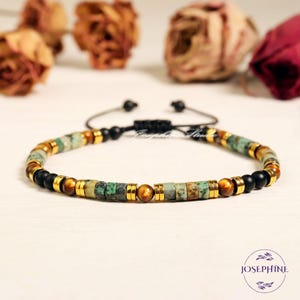May include: A beaded bracelet featuring a mix of natural stone beads in green, brown, and black, accented with gold-colored metal spacers. The bracelet has an adjustable black cord closure. The brand name "Josephine" is visible.