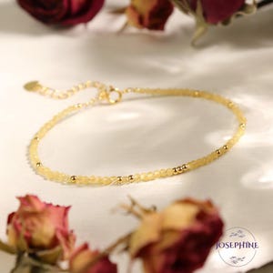 May include: A delicate bracelet featuring small, translucent pale yellow beads interspersed with tiny gold-toned metal spheres. The piece includes a gold-toned metal clasp and an adjustable chain extension. The text JOSEPHINE is visible.