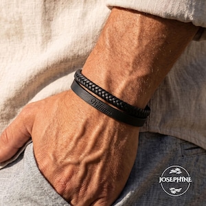 Op de afbeelding: Twee zwarte leren armbanden, één gevlochten en één plat, met de naam "William" gegraveerd, worden op een pols getoond. De persoon draagt een lichtgekleurd overhemd. Een stijlvol accessoire.