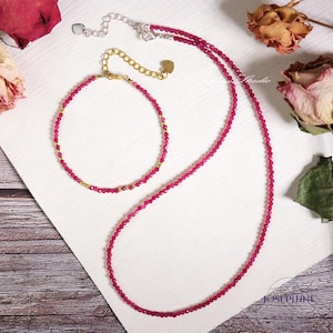 Puede incluir: Un conjunto de dos collares de piedras preciosas rosas, uno largo y otro corto, con detalles dorados. Los collares están hechos de pequeñas cuentas redondas y se muestran sobre una superficie blanca.