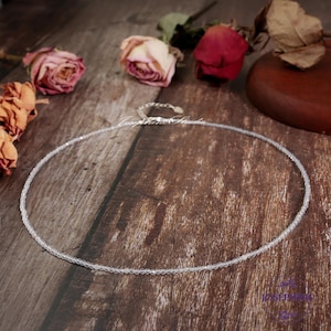 May include: A delicate silver necklace with a chain of clear faceted beads. The necklace is displayed on a wooden surface with dried roses.