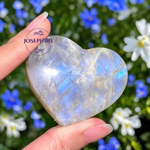May include: A heart-shaped, polished gemstone with a blue and white iridescent sheen. The stone is held between fingers, with the word "JOSEPHINE" printed on the stone. The background features blurred blue and white flowers.