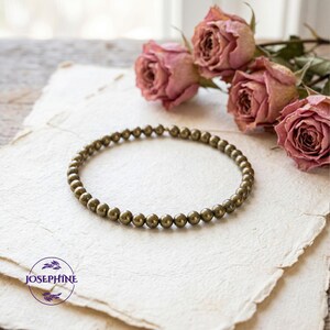 K&ouml;nnte beinhalten: Ein Armband aus kleinen, runden, metallischen Perlen. Das Schmuckst&uuml;ck wird auf einer strukturierten, cremefarbenen Oberfl&auml;che pr&auml;sentiert, mit getrockneten rosa Rosen im Hintergrund. Es ist ein Schmuckst&uuml;ck.
