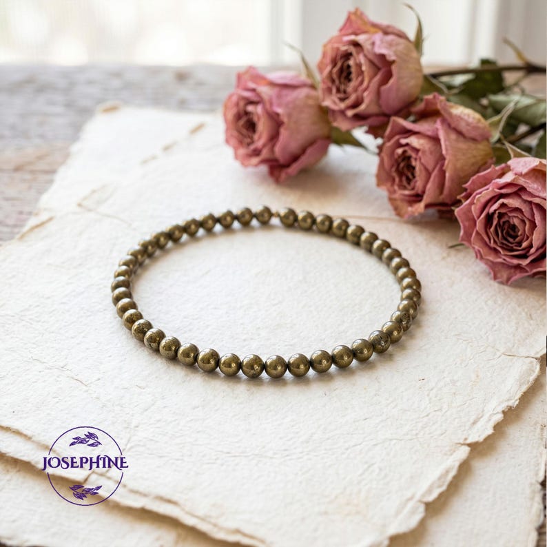 K&ouml;nnte beinhalten: Ein Armband aus kleinen, runden, metallischen Perlen. Das Schmuckst&uuml;ck wird auf einer strukturierten, cremefarbenen Oberfl&auml;che pr&auml;sentiert, mit getrockneten rosa Rosen im Hintergrund. Es ist ein Schmuckst&uuml;ck.