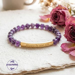May include: A beaded bracelet featuring amethyst stones and a gold-colored bar engraved with the phrase "Always by your side." The bracelet is displayed with dried roses and the brand logo.