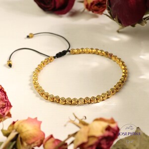 May include: A gold beaded bracelet with a black cord closure. The beads are small and have a geometric shape with a raised design.