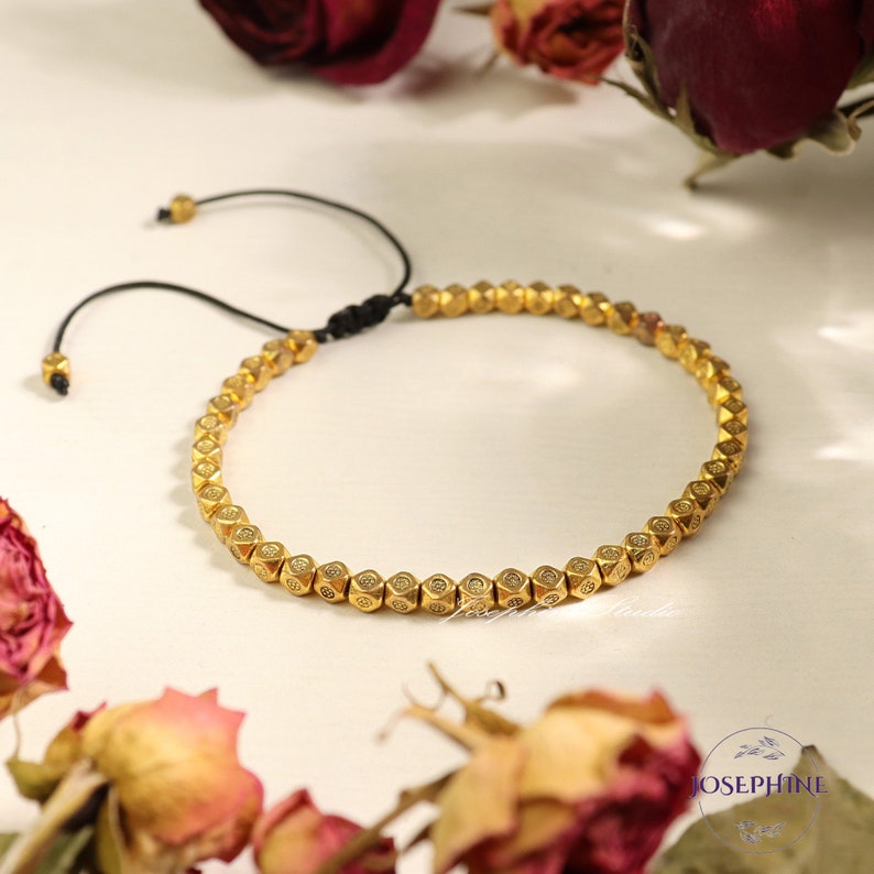 May include: A gold beaded bracelet with a black cord closure. The beads are small and have a geometric shape with a raised design.