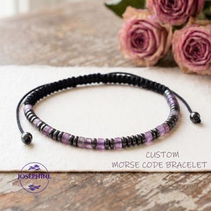 May include: A custom Morse code bracelet with a black woven band and adjustable closure. The bracelet features a combination of small, dark gray and light purple beads. The text "CUSTOM MORSE CODE BRACELET" is visible in the image.