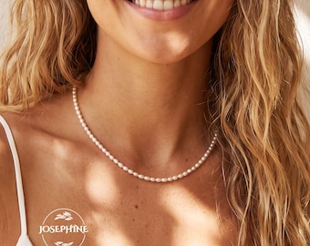 May include: A delicate necklace featuring a single strand of small, uniform white pearls. The necklace rests against the neck, complemented by a pair of simple gold hoop earrings. A circular logo with the text JOSEPHINE is visible in the corner.