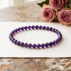 May include: A beaded bracelet made of small, round, deep purple amethyst stones. The bracelet is resting on a cream-colored surface, with dried pink roses in the background. The word "JOSEPHINE" is printed on the bottom right.