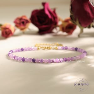 May include: A delicate purple amethyst beaded bracelet with a gold clasp. The bracelet is on a white surface with dried roses in the background.
