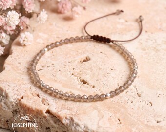 May include: A delicate bracelet featuring small, faceted, translucent gray beads and a single silver-toned metal bead. The bracelet includes a dark brown cord with an adjustable sliding knot closure and the text JOSEPHINE printed in white.