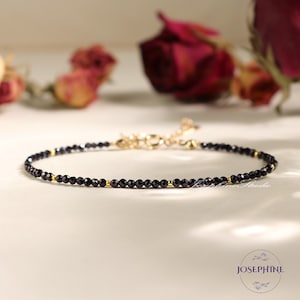 May include: A delicate black and gold beaded bracelet with a clasp closure. The bracelet is made with small, round black beads and gold accents.