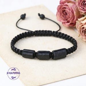 May include: Black macrame bracelet featuring three rectangular black stones. The bracelet has an adjustable sliding knot closure. The word "JOSEPHINE" is in a circle in the lower left corner. Dried pink roses are in the background.
