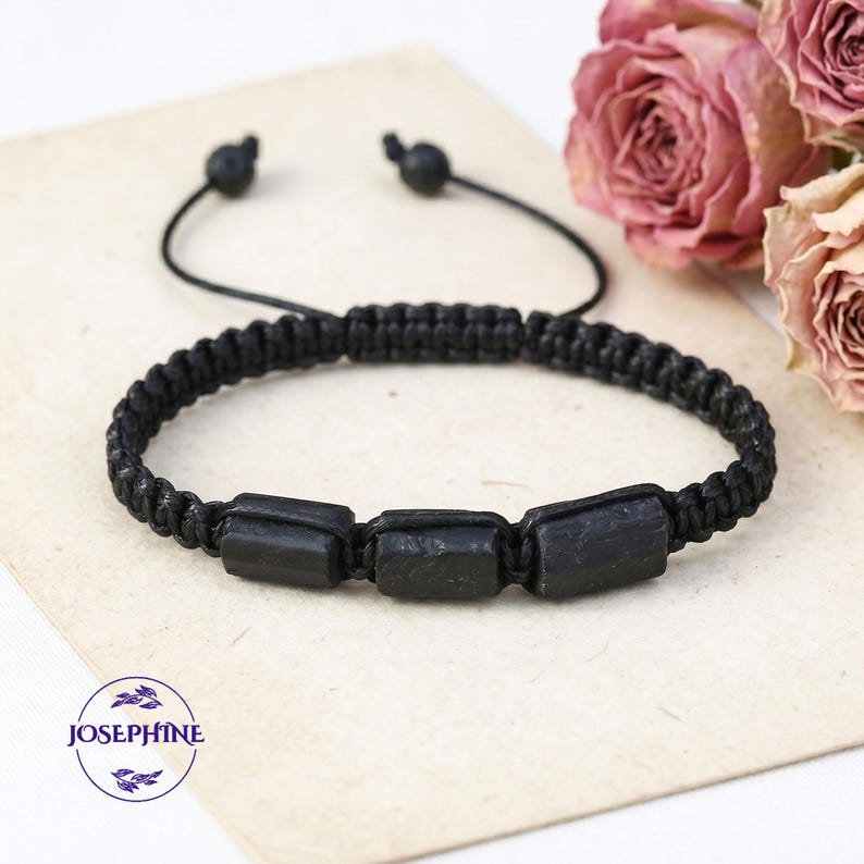 May include: Black macrame bracelet featuring three rectangular black stones. The bracelet has an adjustable sliding knot closure. The word "JOSEPHINE" is in a circle in the lower left corner. Dried pink roses are in the background.