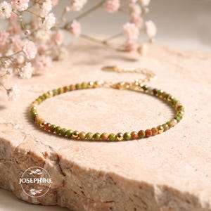 May include: A delicate bracelet featuring small, faceted green and reddish-brown stone beads interspersed with tiny, polished gold-tone metal spheres. The bracelet includes a gold-tone chain extension with a small circular logo tag.