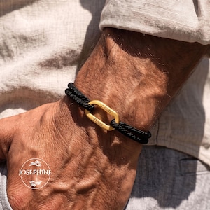 May include: A double-strand black cord bracelet featuring a central gold-toned oval metal link. The link is engraved with the names Lucas and Fiona. A circular logo with the text JOSEPHINE and leaf illustrations is visible in the lower left corner.