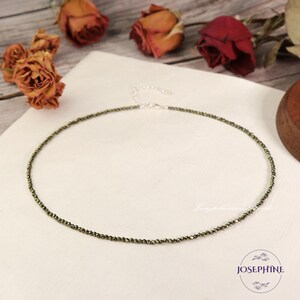 May include: A delicate gold necklace made of small, round pyrite beads. The necklace is on a white surface with dried roses in the background.