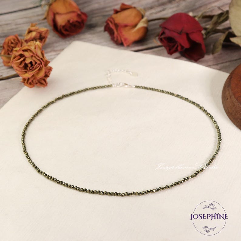 May include: A delicate gold necklace made of small, round pyrite beads. The necklace is on a white surface with dried roses in the background.