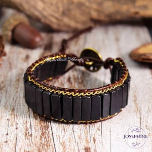May include: A handcrafted bracelet featuring rectangular black beads, brown leather, and gold-colored wire accents. The bracelet has a button clasp and is displayed on a wooden surface. The brand name "Josephine" is visible.