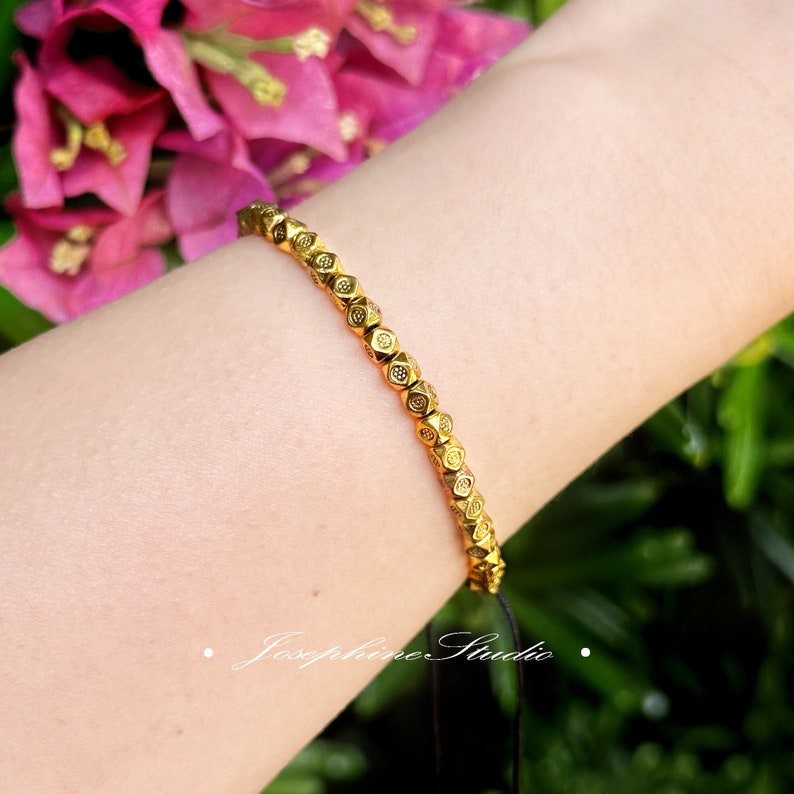 May include: A gold bracelet with a beaded design. The beads are small and have a geometric shape. The bracelet is on a person's wrist.