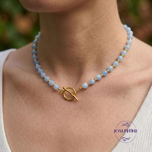 May include: A beaded necklace featuring light blue gemstones and gold-colored accents. The necklace has a toggle clasp and is displayed on a person. The word "JOSEPHINE" is printed in the lower right corner.