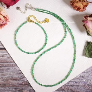 Puede incluir: Un delicado conjunto de joyas con collar y pulsera. El collar y la pulsera están hechos de pequeñas cuentas verdes redondas. El collar tiene un cierre plateado y un dije de corazón dorado. La pulsera tiene un cierre dorado. La palabra "Josephine" es visible.