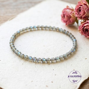 May include: A delicate bracelet made of small, round, gray labradorite beads. The beads have iridescent blue and green flashes. The bracelet is displayed on a textured, off-white surface, with dried pink roses in the background. The brand name "JOSEPHINE" is in a circle.