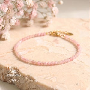 Op de afbeelding: Een delicate armband met kleine, gefacetteerde lichtroze edelsteenkralen in een cirkelvormig patroon. De armband is voorzien van een goudkleurige metalen sluiting en een klein rond label met de merknaam JOSEPHINE in het wit.