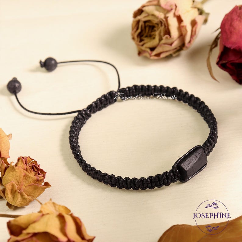 May include: Black braided bracelet with an adjustable sliding knot closure. The bracelet features a rectangular black accent piece. The background includes dried roses and the word "Josephine" in a logo.