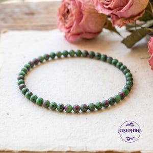 May include: A beaded bracelet featuring small, round beads in shades of green and red. The bracelet is displayed on a cream-colored surface, with dried pink roses in the background. The brand name "JOSEPHINE" is visible.