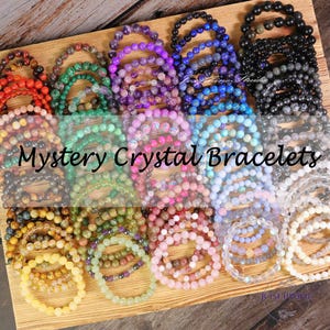 May include: A collection of colorful crystal bracelets, each with a different stone and color. The bracelets are arranged in rows on a wooden surface. The text "Mystery Crystal Bracelets" is displayed across the image.
