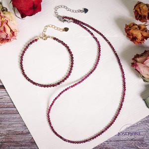 May include: A set of two necklaces made with small, dark red gemstone beads. The necklace is longer than the bracelet. Both pieces are on a white surface.